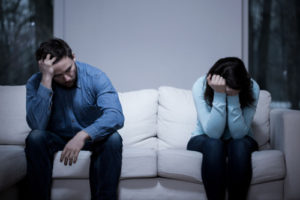 Photo of a couple sitting at opposite ends of a couch and holding their heads in resentment and despair.